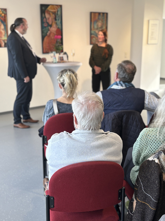 Zwei MEnschen unterhalten sich im Hintergrund; im Vordergrund sitzten Menschen auf Stühlen